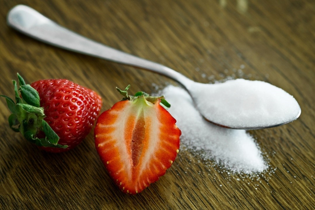Spoonful of sugar next to a strawberry that has been cut in half vertically on a wooden surface.